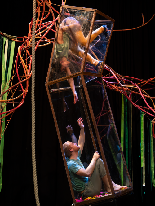 2 performers in a glass and metal structure on a stage, one upside down, reaching for each other. The Hideout